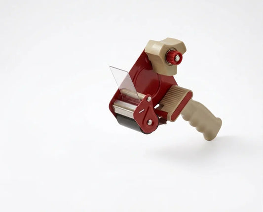 A professional studio photograph of a red and beige handheld industrial tape gun dispenser, shown from a three-quarter angle against a seamless white background. The device features a prominent red metal frame, a clear plastic tape guide shield, a multi-roller application head with a serrated cutting blade, a large tan-colored plastic core holder with a red tension knob, and a matching textured tan ergonomic handle grip. It is posed in an empty, ready-to-be-loaded state.